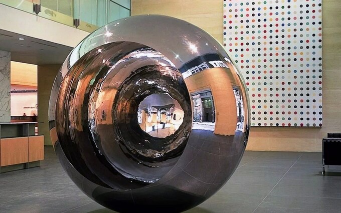 The image shows a spacious lobby with modern art installations. Dominating the foreground is a large, highly polished metallic sphere with a tunnel-like hole through its center, reflecting the surrounding environment in its shiny surface. On the right, there is a large canvas covered with a grid of colorful dots, evoking a playful yet orderly aesthetic typical of modern art. The backdrop features neutral-colored walls and a light-colored floor, with a reception desk visible in the upper left corner, creating a welcoming yet sophisticated ambiance. A black seating area to the far right adds to the functional aspect of the space. The overall impression is one of a contemporary and artistic venue designed to intrigue and engage viewers.