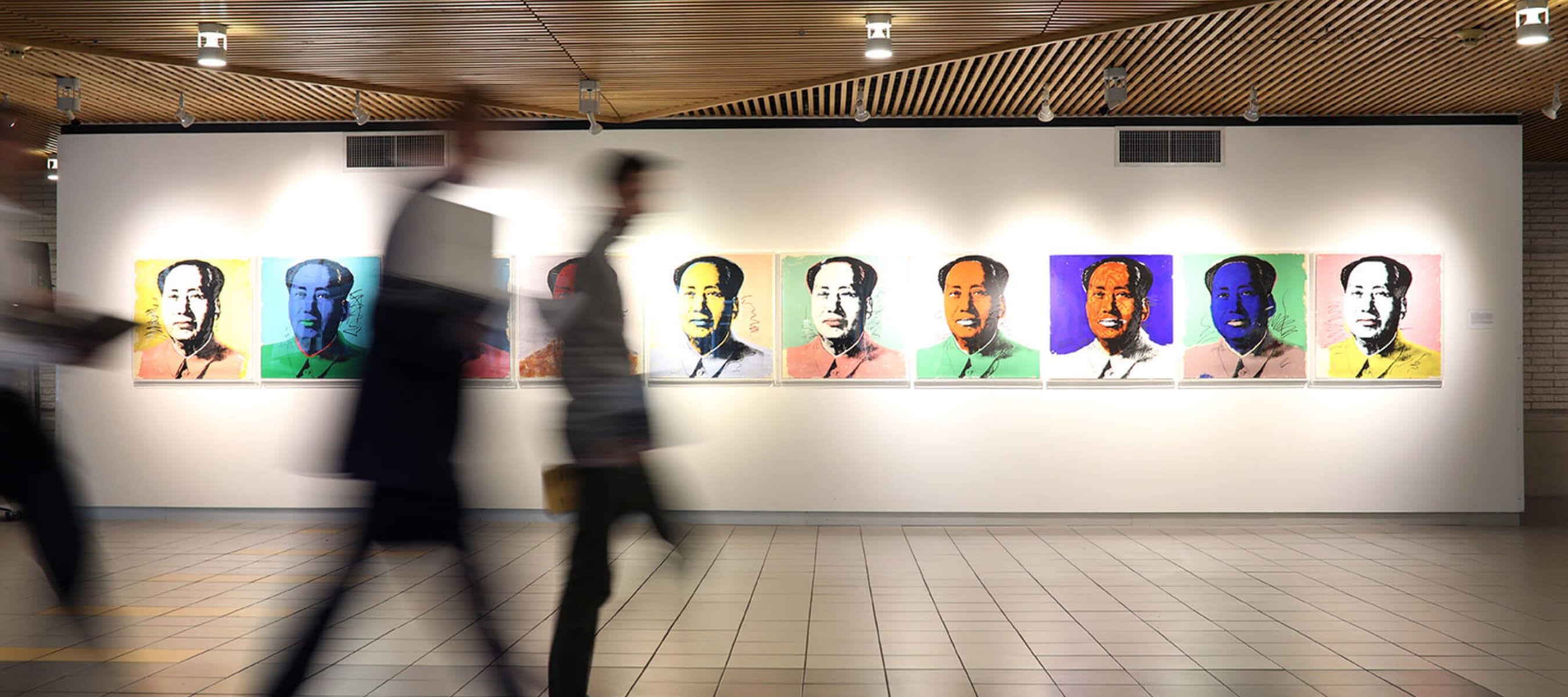In an art gallery, eight pop art-style portraits of the same figure with different colored backgrounds are displayed on a white wall. Blurred figures of visitors pass by in the foreground, suggesting movement through the gallery. The portraits are illuminated by track lighting from the wood-paneled ceiling above the tiled floor.
